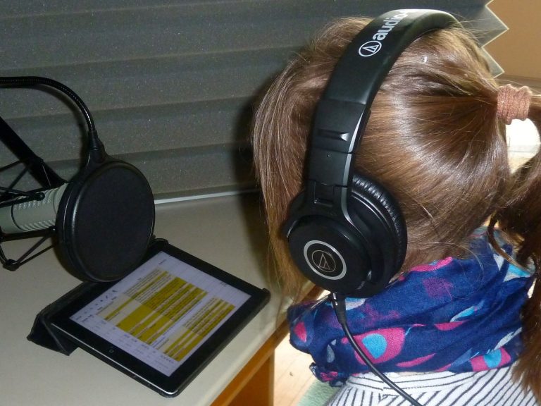 Ein Mädchen sitzt mit Kopfhörern vor einem Tablet und einem Aufnahmemikrophon.